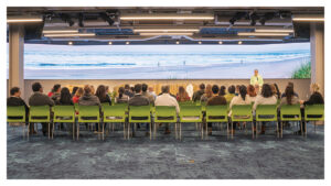 Photograph of the Beach @ The Cove. Green chairs set up with a large group of people attending the presentation. Wide screen displaying an ocean and skyline.